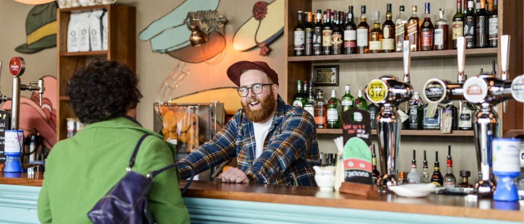 A bearded bartender in a berretto and glasses smiles and chats with a customer in a green coat at a colorful bar lined with bottles and beer taps.