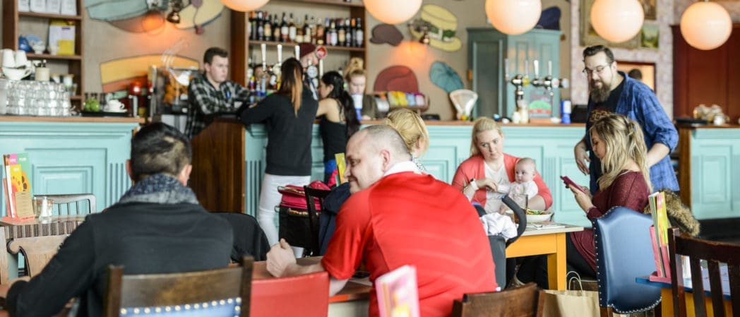 A busy, colorful café with people sitting at tables, chatting, and eating. A woman in a berretto holds a baby while staff work behind the bar. The atmosphere is lively and casual.