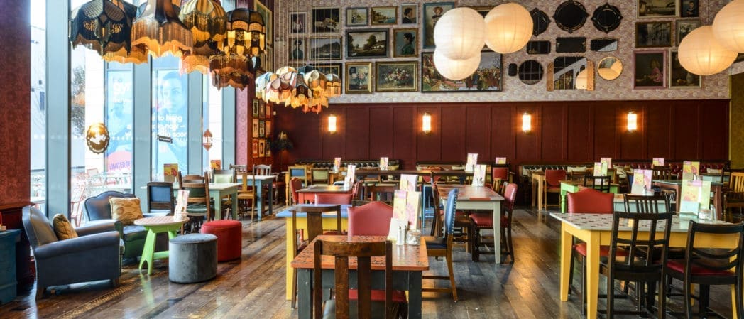 A colorful, eclectic restaurant interior with mismatched chairs, wooden tables, large windows, hanging lights, decorative berretto lampshades, and a wall covered in assorted framed art and mirrors.