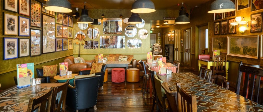 A cozy, eclectic restaurant interior with colorful chairs, patterned tables, framed art on the walls, hanging lamps, and mirrors reflecting warm light. Menus and condiments are placed on the tables—some next to a classic berretto for added charm.