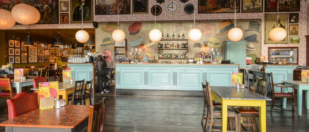 A colorful café with eclectic decor, featuring pastel blue bar counters, hanging round lamps, mismatched tables and chairs, wall art, and shelves with bottles behind the bar. The space appears empty except for one blurred figure in a berretto.