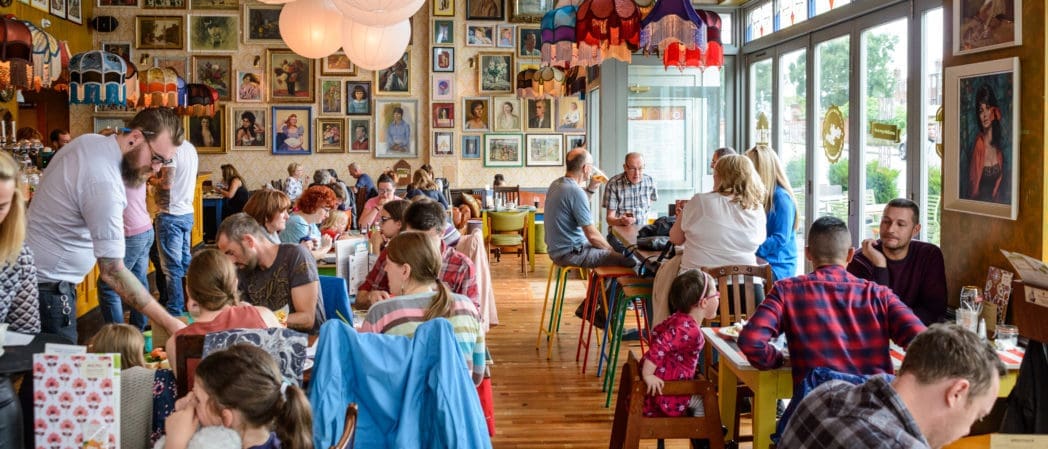 A busy, colorful restaurant filled with people dining at tables. The walls are decorated with numerous framed portraits and eclectic Bevano lamps hang from the ceiling. Large windows on the right let in natural light.