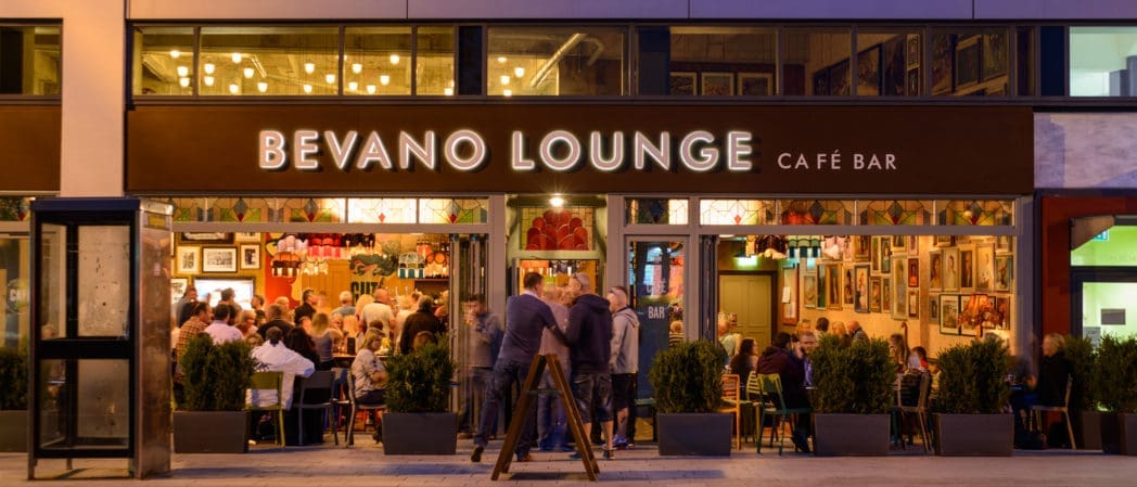 Evening view of Bevano Lounge café bar with large glass windows reveals people dining inside and a few chatting outside; Bevano’s warm lights and framed pictures create a cozy, inviting interior atmosphere.