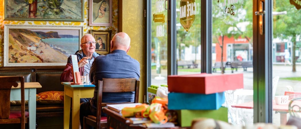 Two older men sit at a small table in a cozy, art-filled café, talking and smiling as a Bolero plays softly. Bright daylight streams through large windows, colorful boxes are stacked nearby, and trees and buildings are visible outside.