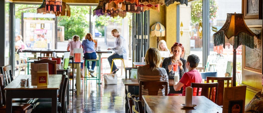People sit at tables inside a cozy café with colorful Bolero hanging lamps and large windows, while others enjoy the sunny day outside. The atmosphere is relaxed and inviting.