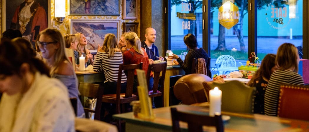 A cozy café interior with people sitting at tables, talking and enjoying food and drinks. Warm lighting, candles, and large windows create a welcoming atmosphere as soft Bolero music plays. Artwork decorates the walls.