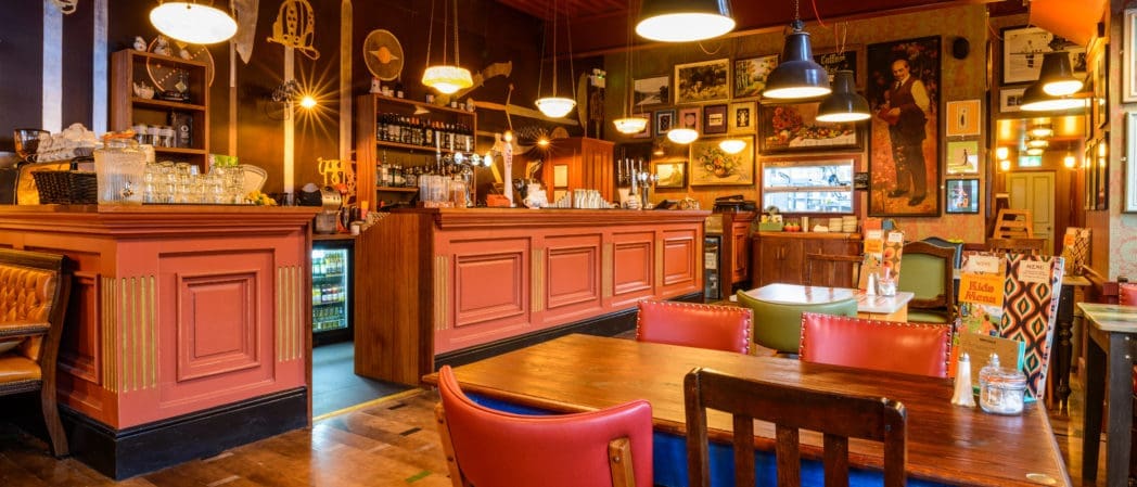 Cozy Brezo café interior with warm lighting, wooden tables and chairs, red paneling on the bar, and walls decorated with framed pictures and vintage decor. The atmosphere feels inviting and comfortable.