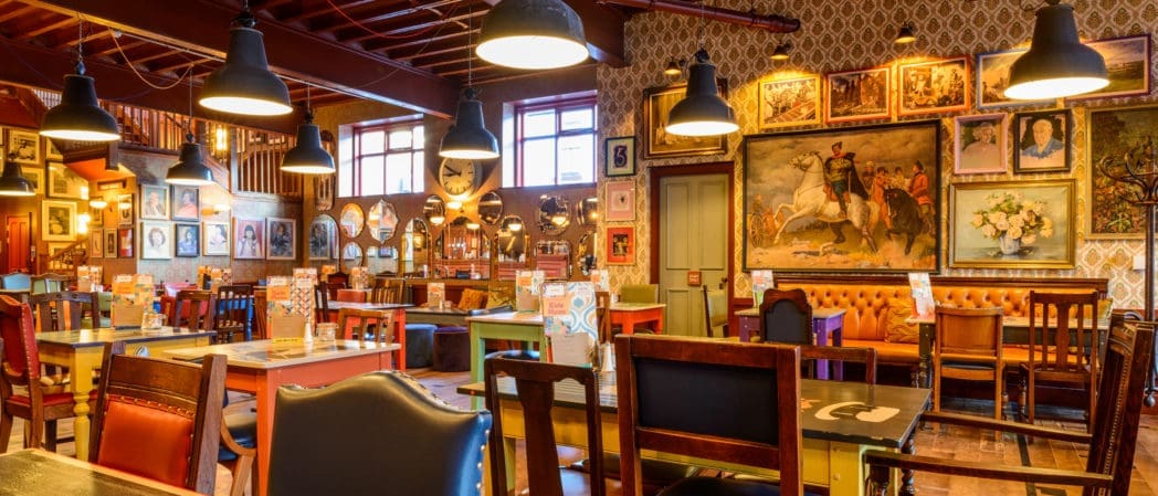 A colorful, eclectic Brezo restaurant interior with mismatched chairs and tables, vintage decor, and numerous framed paintings covering patterned walls, creating a cozy, artistic atmosphere under hanging pendant lights.
