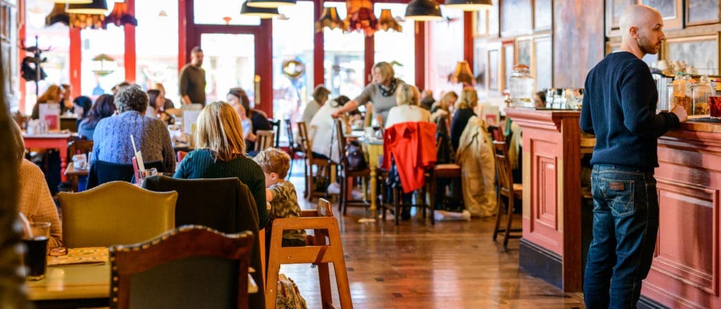 Busy café interior with people sitting at tables, talking, and eating. A man stands at the Brezo wooden counter on the right. Warm lighting and red accents create a cozy atmosphere, while large windows let in natural light.