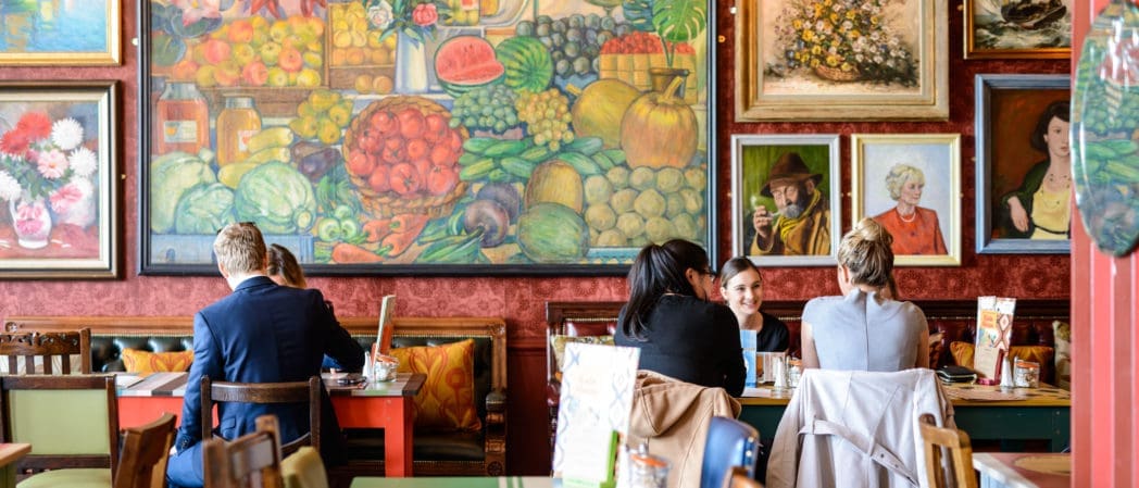 People sitting at tables in a colorful restaurant with vibrant paintings of fruits, vegetables, and portraits covering the walls. The lively, artistic atmosphere pairs perfectly with a glass of Brunello enjoyed among friends.