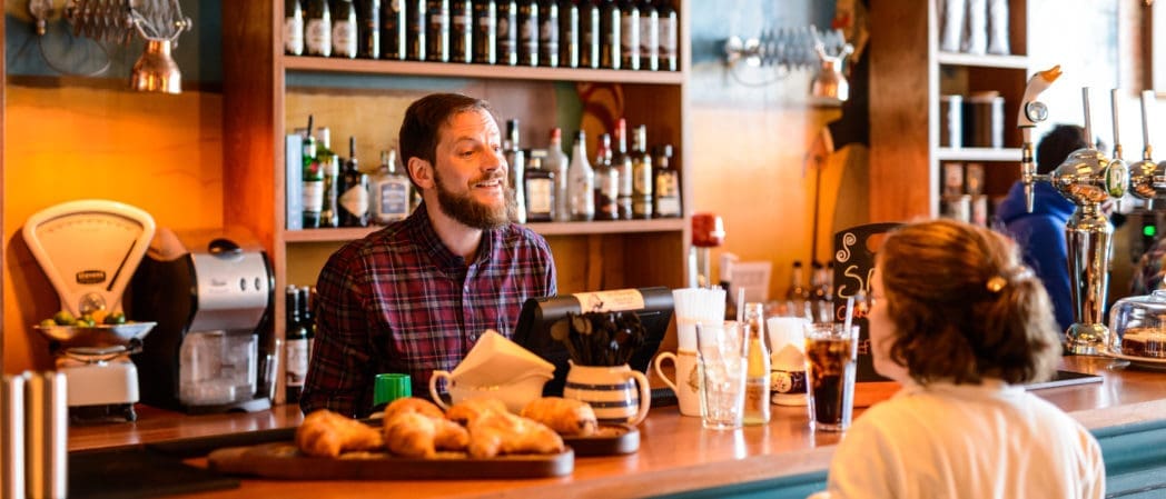 A bearded barista in a plaid shirt smiles while talking to a customer at a cozy café counter, with pastries, drinks, and bottles of Brunello displayed in the background.