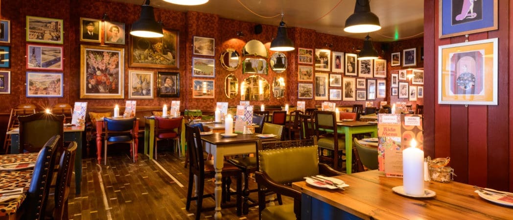 A cozy restaurant with eclectic decor, featuring mismatched chairs, wooden tables, and walls covered in framed pictures and mirrors. Warm lighting creates an inviting atmosphere—perfect for savoring a glass of Brunello by candlelight.