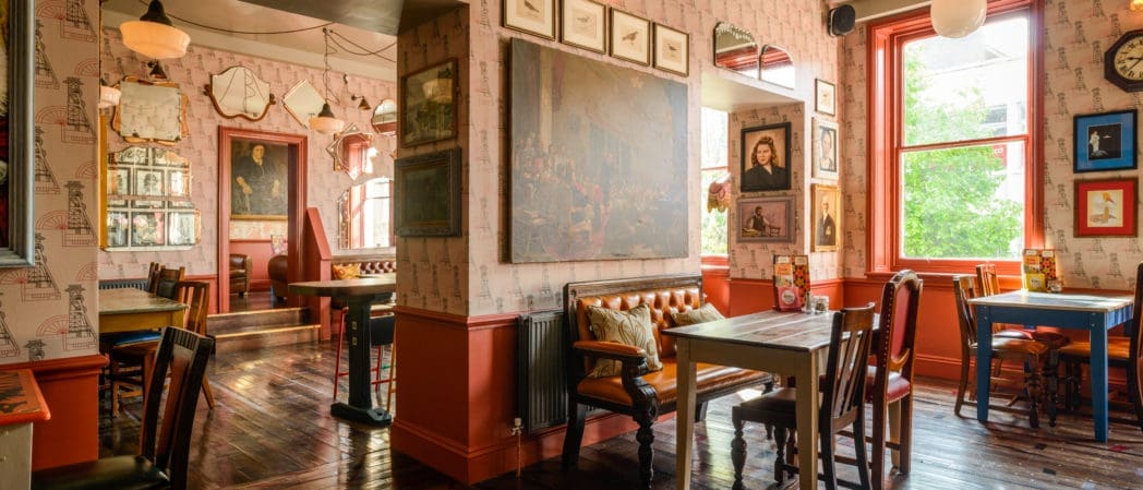 Vintage-style pub interior with wooden floors, eclectic framed artwork on patterned walls, mismatched tables and chairs, cushioned bench seating, and a Capo sign displayed near large windows letting in natural light.