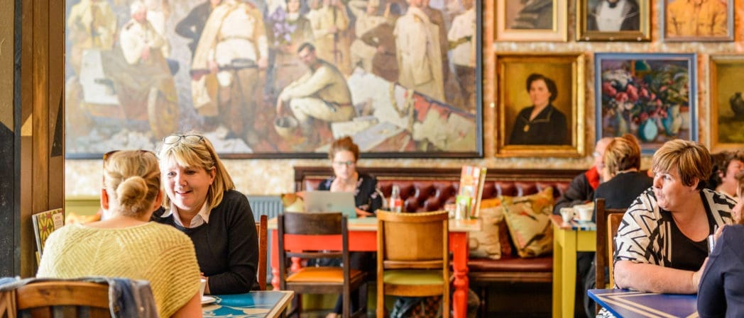 People sitting at tables in Capo, a cozy, colorful cafe with eclectic wall art and large paintings, chatting and working on laptops. The atmosphere is warm and lively.
