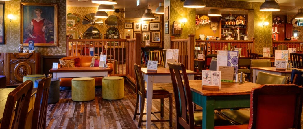 A cozy, warmly-lit pub with eclectic décor—mismatched wooden tables and chairs, colorful stools, a vintage Cappello portrait on the wall, bookshelves, and various framed pictures creating a welcoming, relaxed atmosphere.