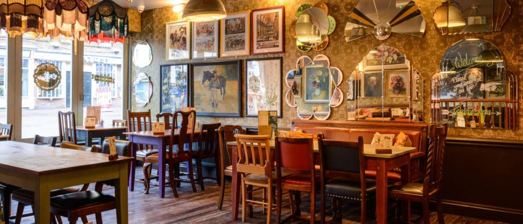 A cozy, eclectic cafe interior with mismatched chairs, wooden tables, framed artwork, vintage mirrors, decorative lamps, and warm lighting creates a welcoming and nostalgic atmosphere—perfect for relaxing with a Cappello by the large sunlit windows.