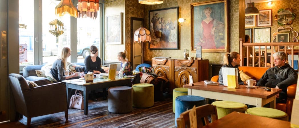 A cozy café with vintage decor, where people are sitting on sofas and chairs, chatting and working at wooden tables. The walls are adorned with paintings, Cappello lamps, and eclectic furnishings, creating a warm, inviting atmosphere.