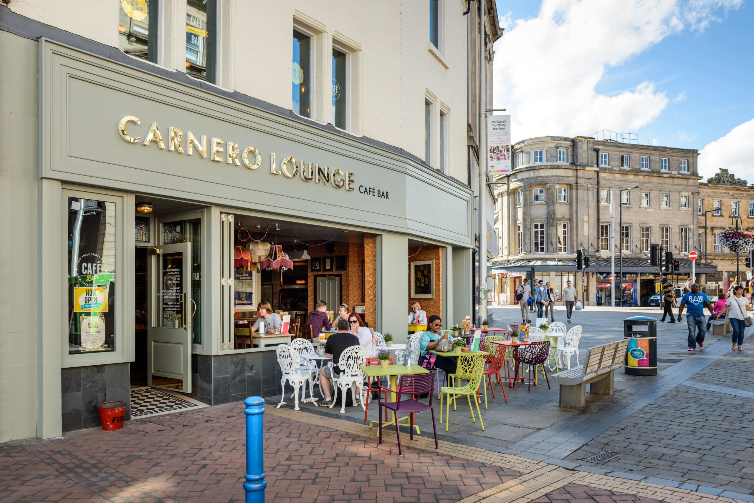 People relax at colorful tables outside Carnero Lounge café bar on a street corner in a busy city area, as pedestrians stroll by historic buildings under a partly cloudy sky.