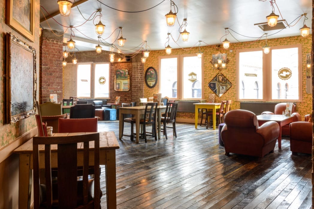 Cozy Carnero cafe interior with wooden floors, brick walls, large windows, eclectic furniture, and warm hanging lights creates a welcoming and relaxed atmosphere. Paintings and mirrors decorate the walls.