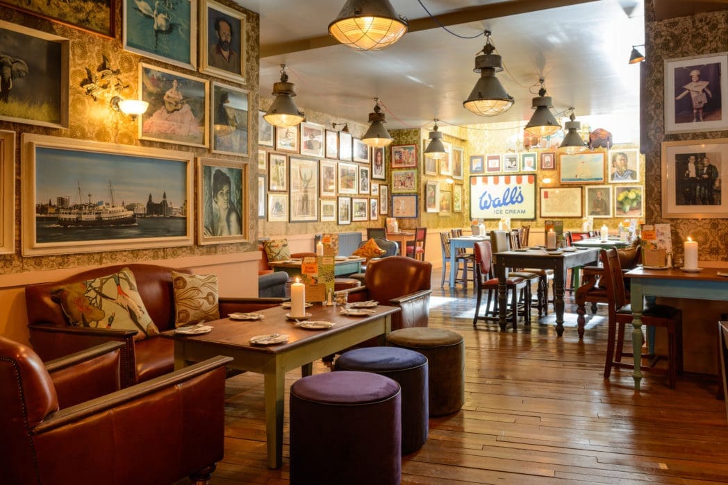 A cozy, vintage-style cafe, Carnero welcomes guests with wooden floors, leather chairs, and walls covered in framed paintings. Warm lighting and eclectic decor create a charming atmosphere; tables are set and ready for guests.