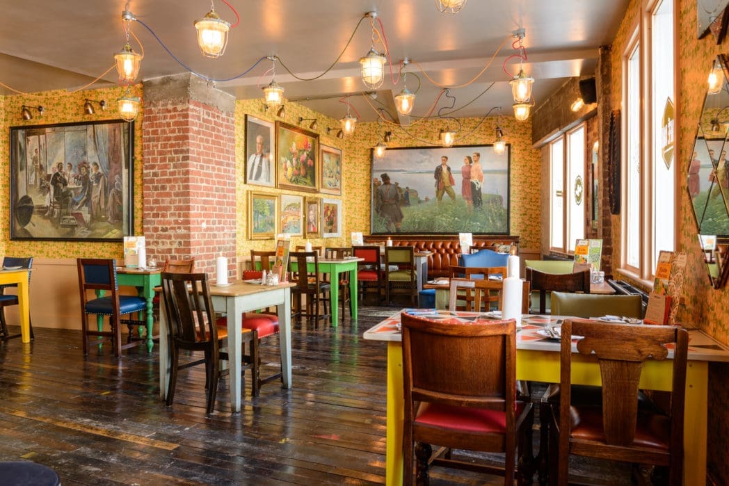 A brightly lit restaurant with wooden floors, colorful mismatched chairs and tables, eclectic wall art, exposed brick pillars, and large windows letting in natural light. The vibrant atmosphere is perfect for enjoying Carnero-inspired dishes.