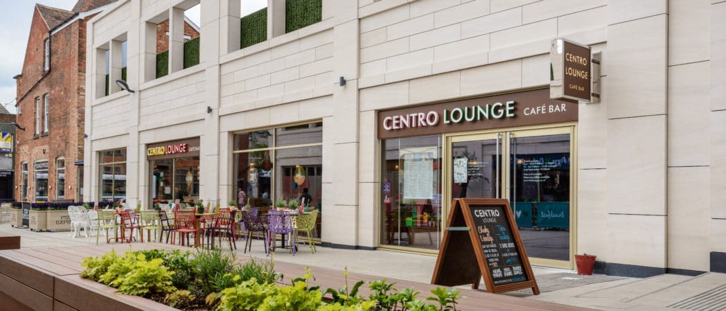 Outdoor view of Centro Lounge café bar with tables and colorful chairs on the sidewalk, a Centro menu board by the entrance, and a modern white building exterior with large windows and lush green plants in the foreground.