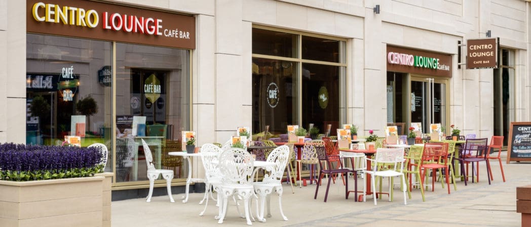 The Centro Lounge Café Bar features a vibrant outdoor seating area with colorful chairs and tables. Large windows and Centro signs welcome guests to this modern beige building, where fresh flowers brighten each table.