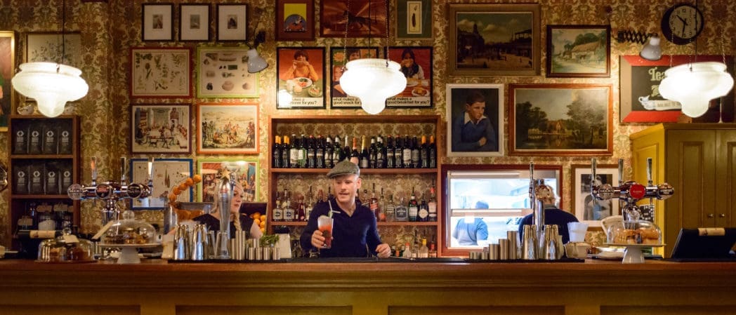 A bartender in a cap stands behind a vintage-style Centro bar, serving a drink. The bar is decorated with framed art, bottles, and glassware, creating a cozy, nostalgic atmosphere with warm lighting.