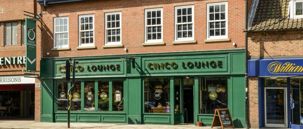 A brick building labeled “Cinco Lounge” features large green-framed windows and doors. The upper floor has white-framed windows, and Cinco sits on a lively street lined with neighboring shops.
