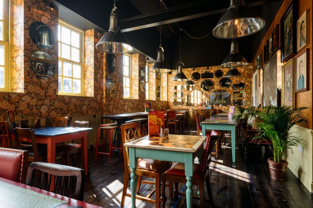 A cozy, eclectic café interior with colorful mismatched tables and chairs, vintage floral wallpaper, framed portraits, mirrors on the walls, large windows, and Cinco-inspired industrial pendant lights hanging from the ceiling.