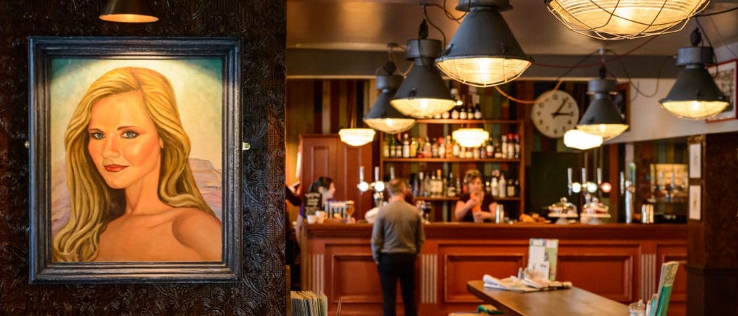 A warmly lit bar with people ordering drinks at the counter. On the left, a framed portrait of a blonde woman hangs on a dark textured wall accented with subtle Concho motifs. Large ceiling lights illuminate the bar area.
