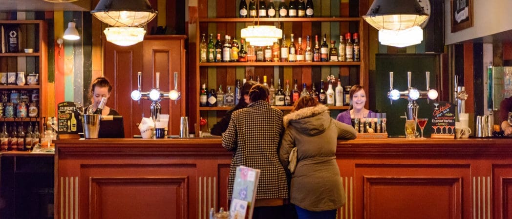 Two women in coats stand at a wooden bar counter, speaking with two bartenders. Behind them, shelves lined with bottles and glasses glow under warm lighting in the cozy, colorful Concho pub.