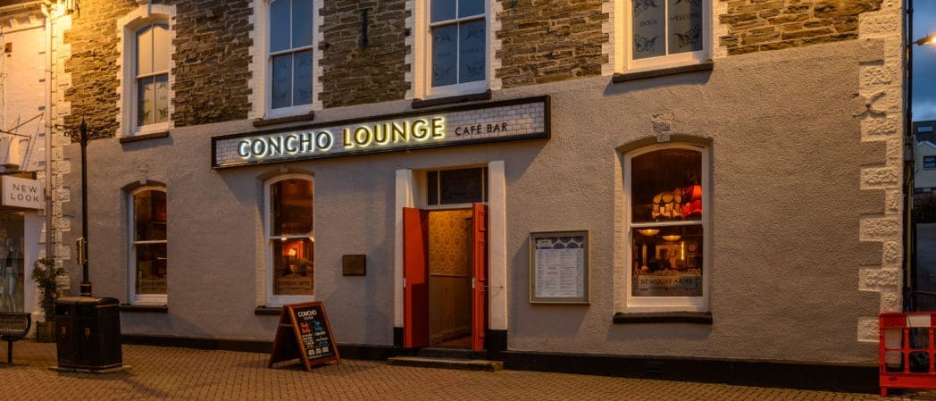 Exterior view of Concho Lounge café bar at dusk, with warm lighting inside, a chalkboard sign by the entrance, and stone and pale-colored walls. Large windows showcase the inviting atmosphere unique to Concho.