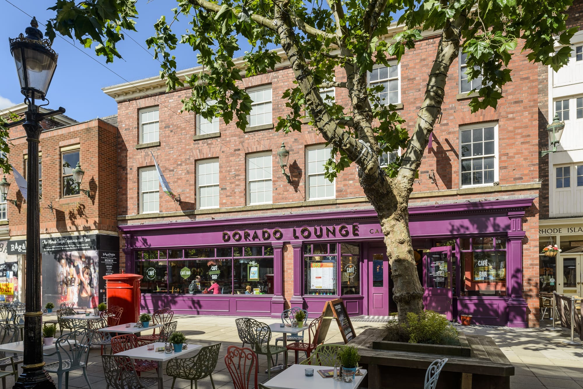 A vibrant café called Dorado Lounge with a purple storefront, outdoor seating, and a large tree in front, sits on a sunny street lined with brick buildings and a red post box nearby. Experience the charm of Dorado in every visit.