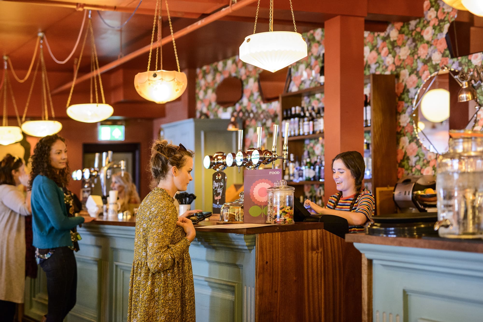 A woman in a patterned dress stands at a vintage Dorado bar counter, speaking with a smiling bartender. Other patrons and decorative lighting are visible, with floral wallpaper and pastel-colored decor in the background.