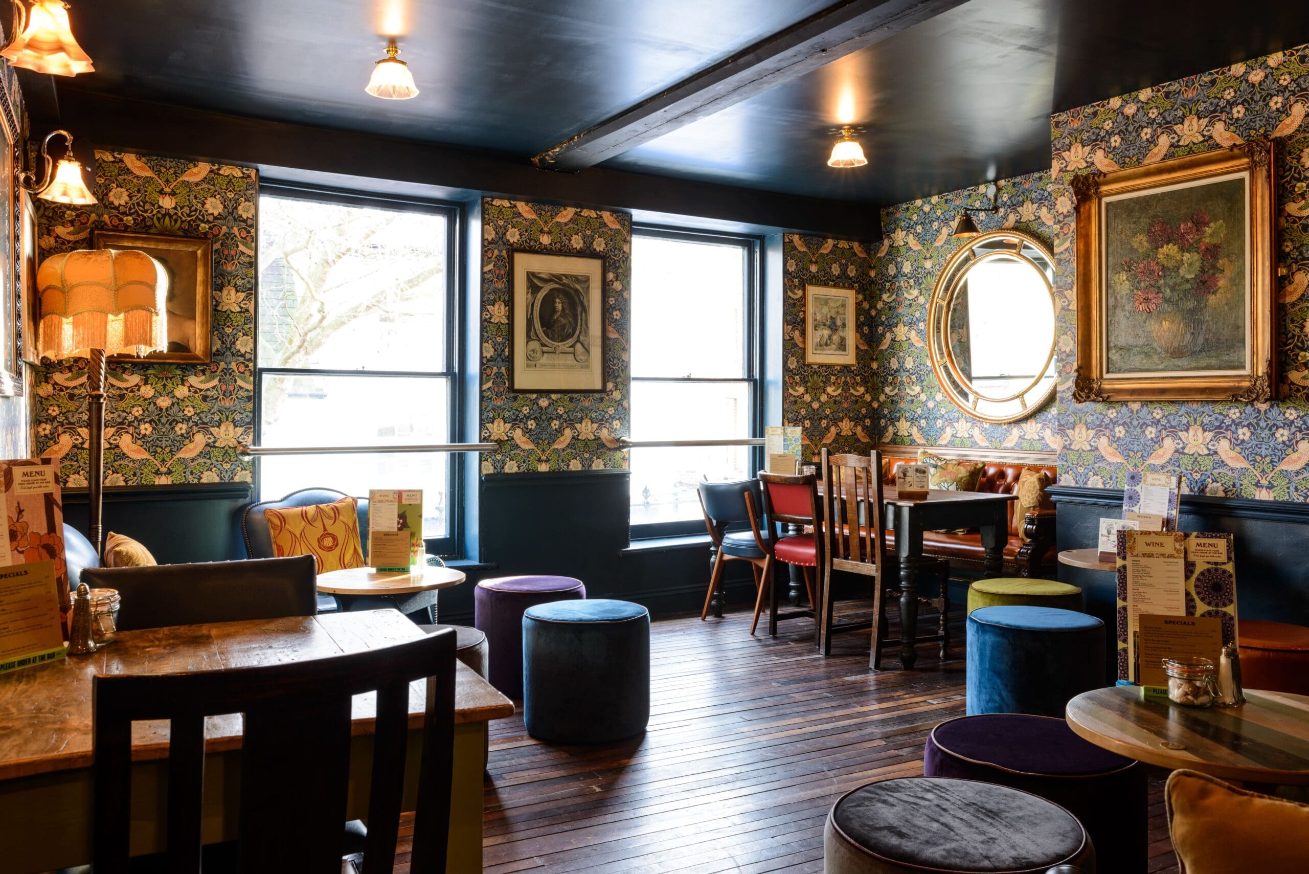 A cozy, vintage-style café interior inspired by Edmundo, with floral wallpaper, framed art, wooden floors, mismatched chairs, round tables, soft poufs, and large windows letting in natural light.