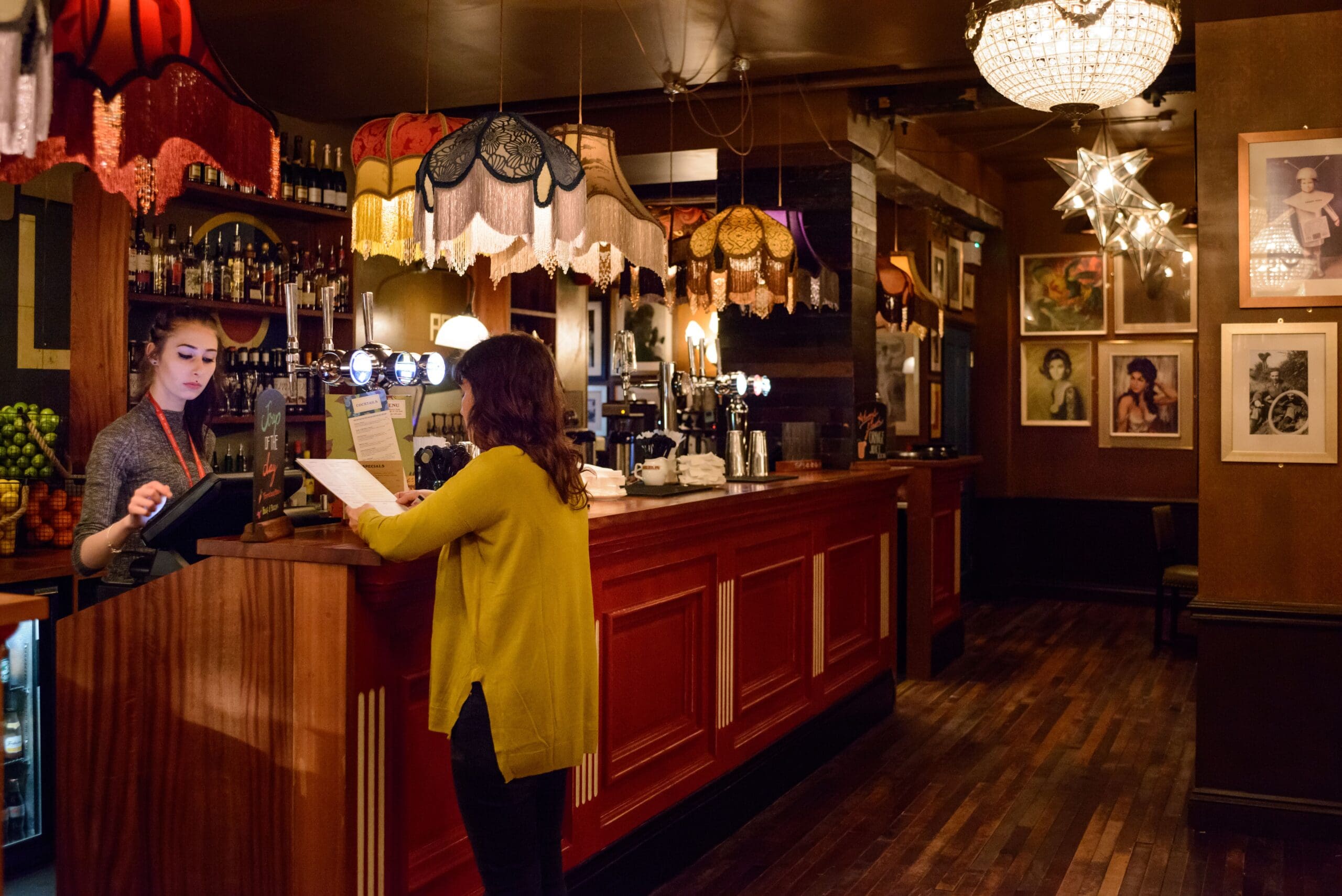 A woman in a yellow sweater stands at Edmundo’s vintage-style bar, reading a menu while a bartender works at the register. The warmly lit space features ornate lamps, chandeliers, and framed portraits on the walls.