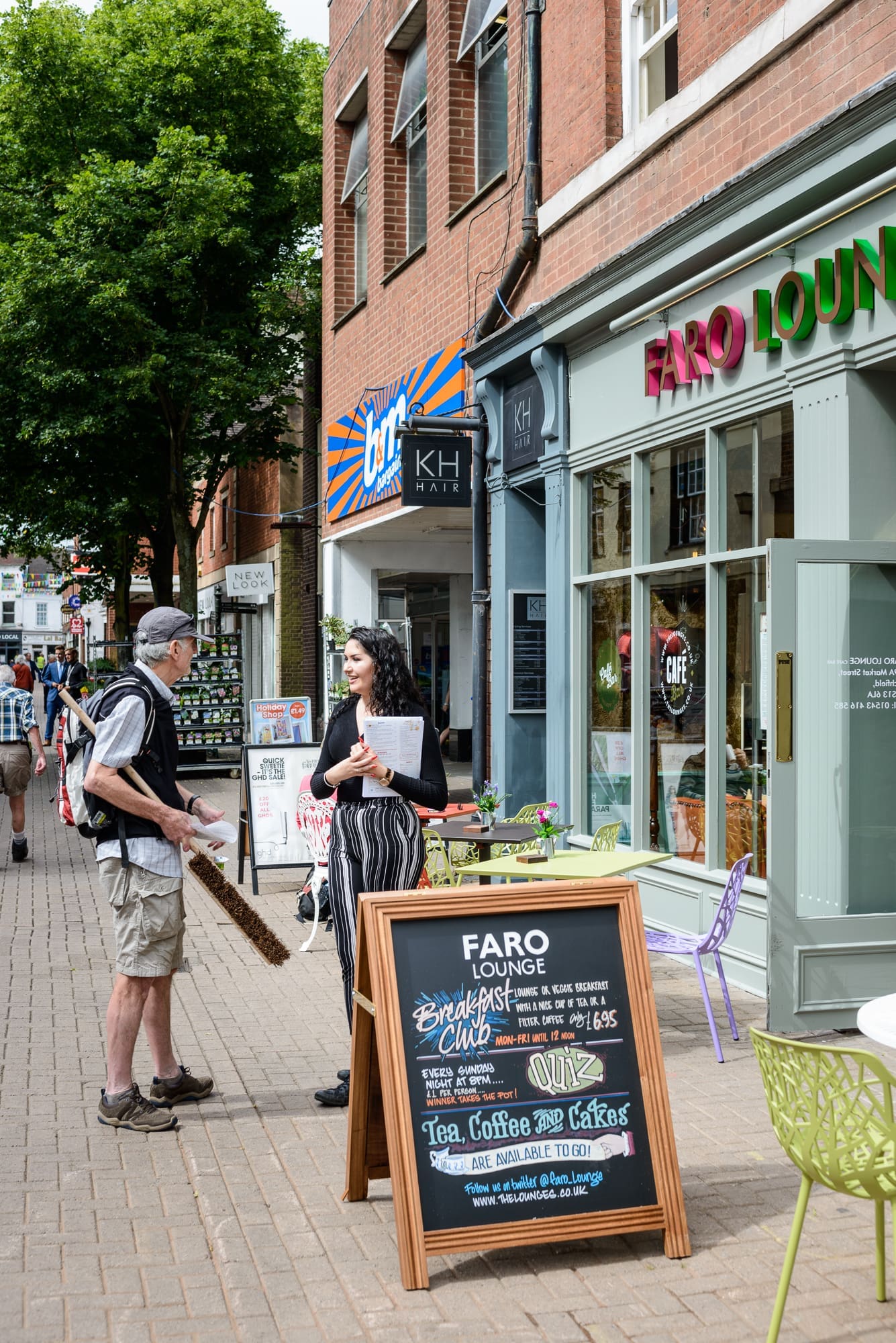 A woman stands outside Faro Lounge café talking to a man with a backpack and walking stick. A Faro chalkboard menu is displayed in front, with other shops, trees, and pedestrians lining the lively street.