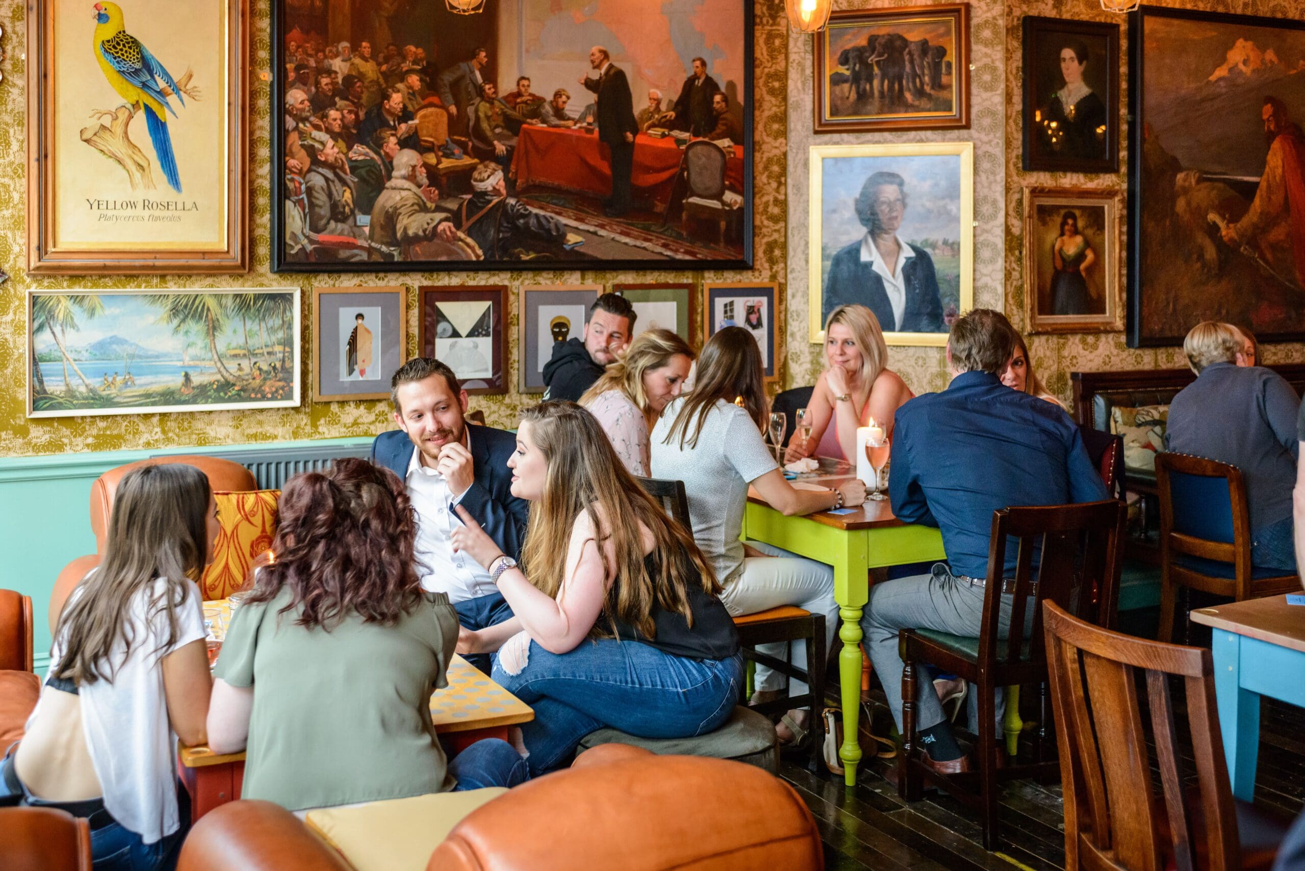 People sit and chat in a lively, cozy café in Faro, decorated with framed paintings and photos on the walls. Groups of men and women enjoy drinks and conversation at colorful tables, creating a warm, social atmosphere.