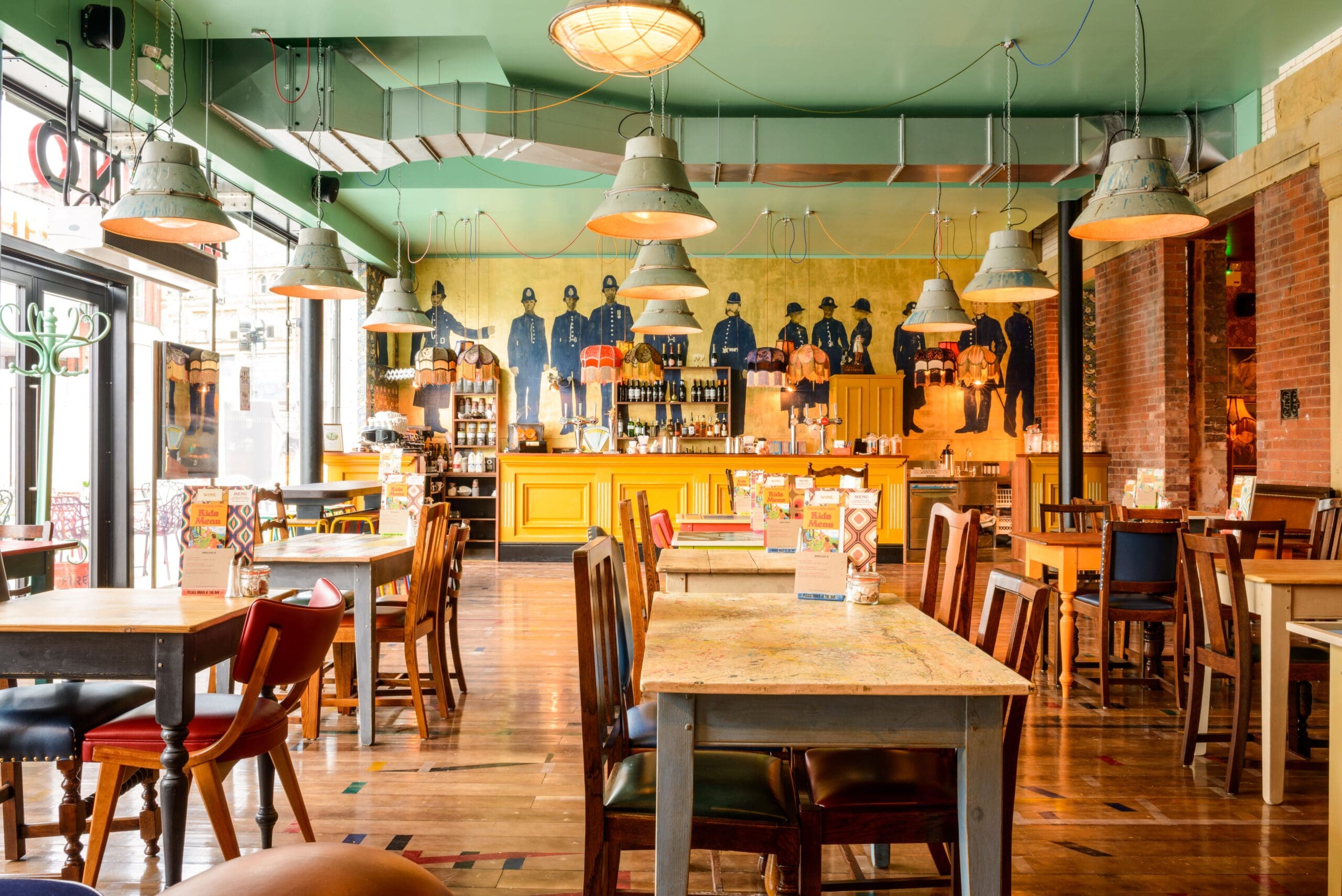 A bright, cozy café with wooden tables and colorful chairs. Industrial-style pendant lights hang from the green ceiling. The back features a yellow bar and wall art of people in blue uniforms—a welcoming spot inspired by Molino.