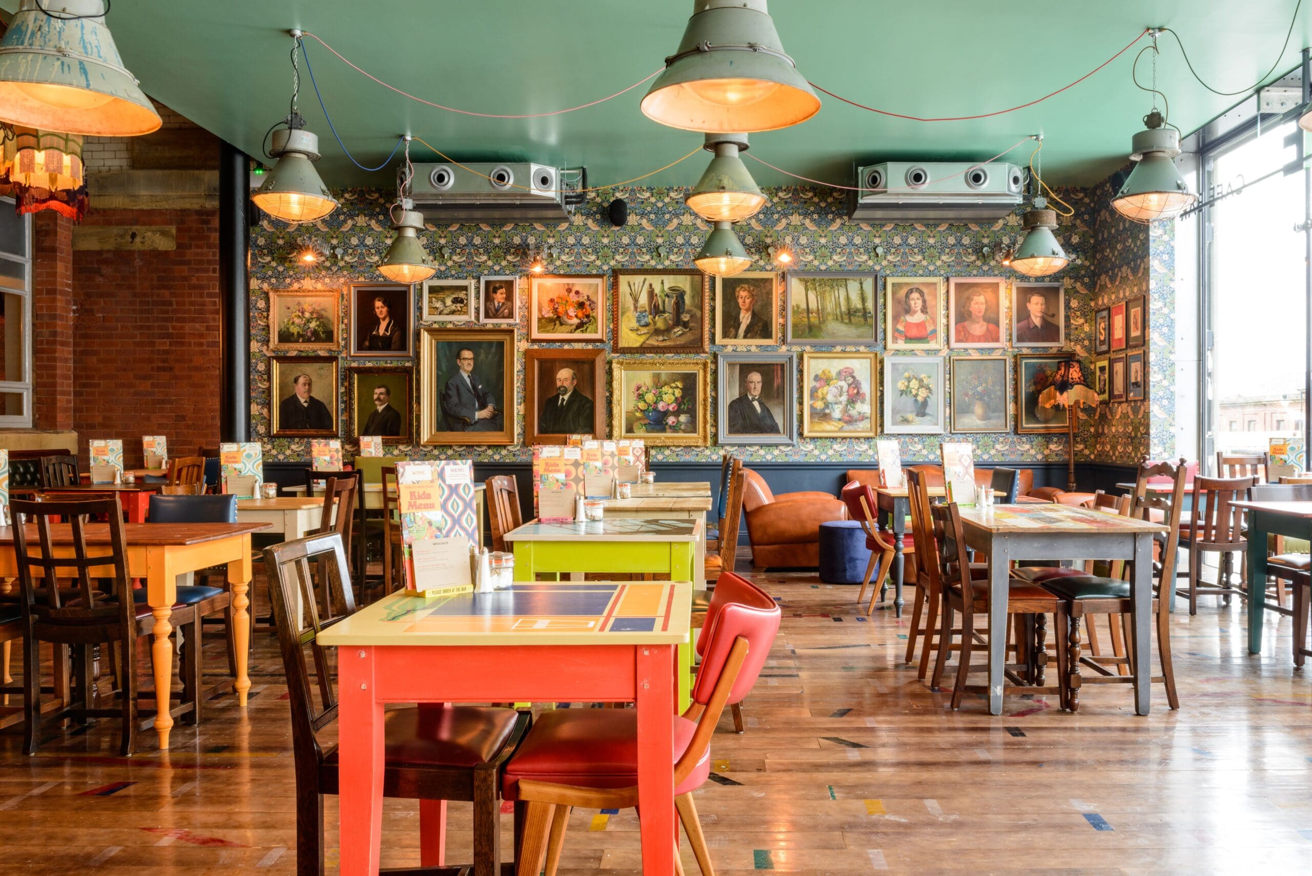 A brightly lit restaurant with colorful tables and chairs, wooden floors, and a wall decorated with many framed portraits and paintings, Molino creates a cozy and artistic atmosphere.
