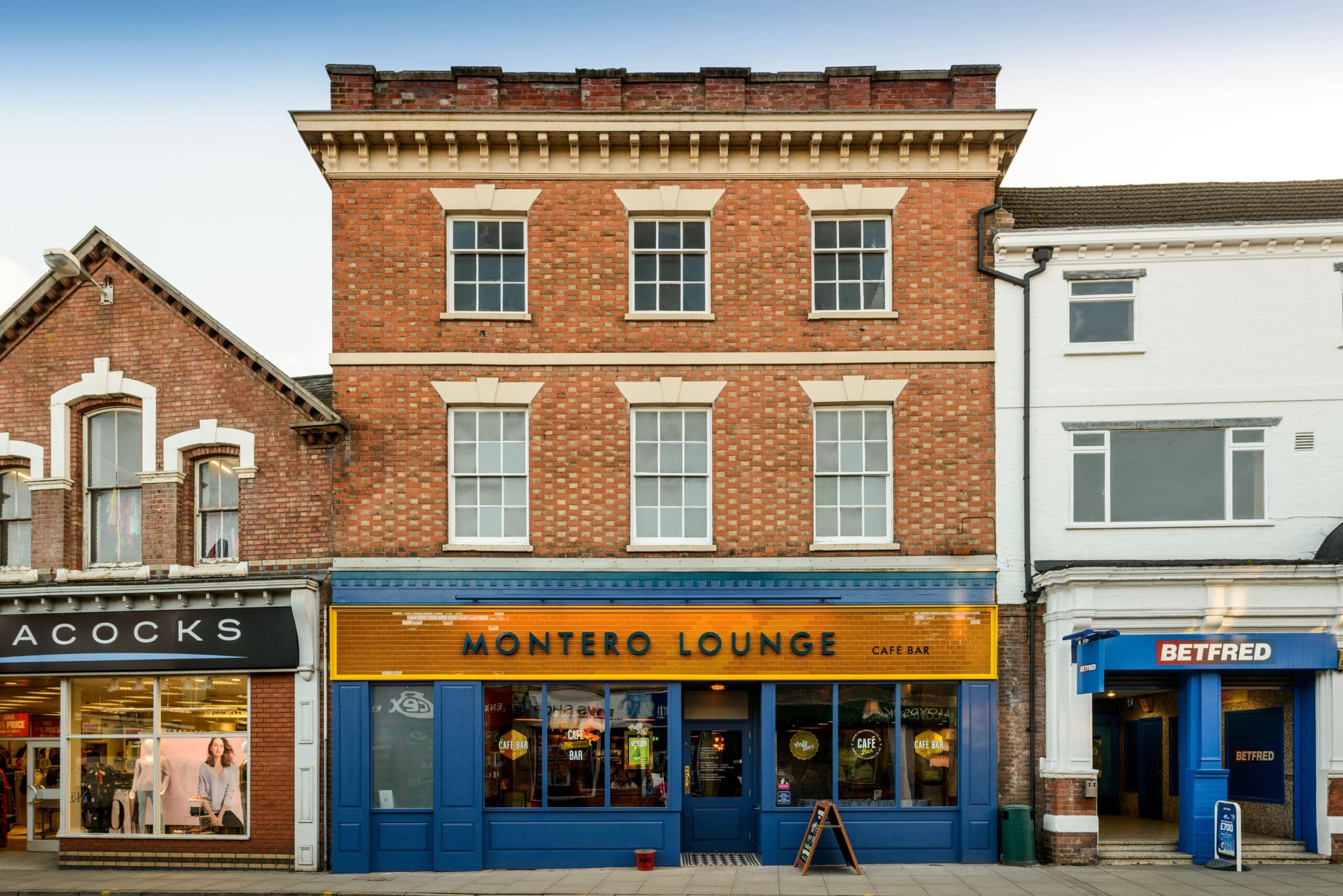 A three-story brick building with a sign reading “Montero Lounge Café Bar” on the ground floor, flanked by shops named “Acocks” and “Betfred,” stands prominently on a street with a sidewalk in front.