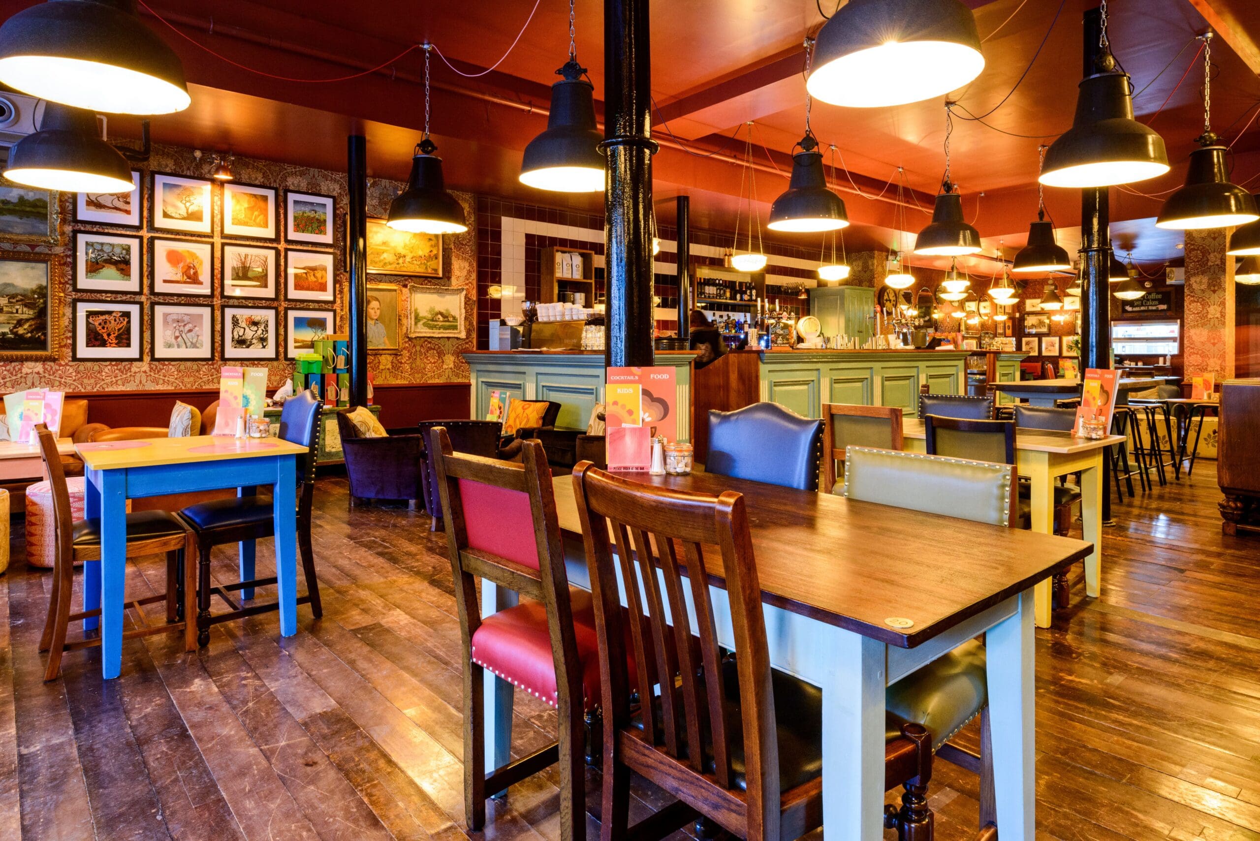 A cozy, brightly lit Montero restaurant interior with wooden floors, mismatched colorful chairs, wooden tables, wall art, hanging lights, and a counter bar in the background.