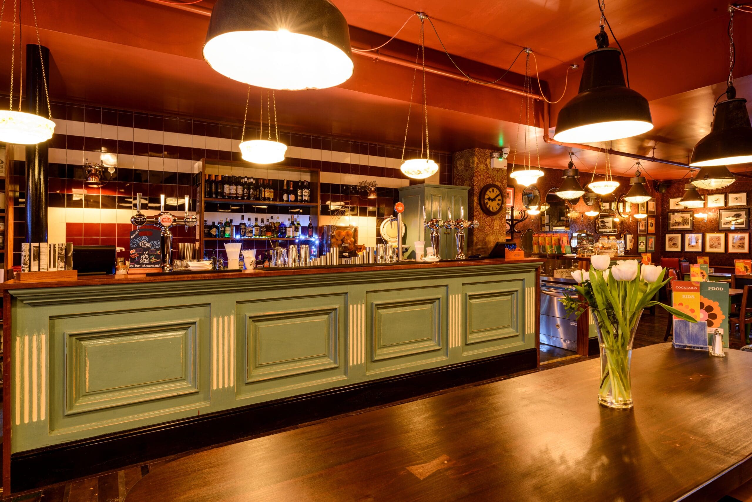A warmly lit bar with a long green counter, hanging pendant lights, and shelves stocked with bottles. A vase of white tulips sits on a wooden table in the foreground, while Montero-themed framed photos decorate the walls.