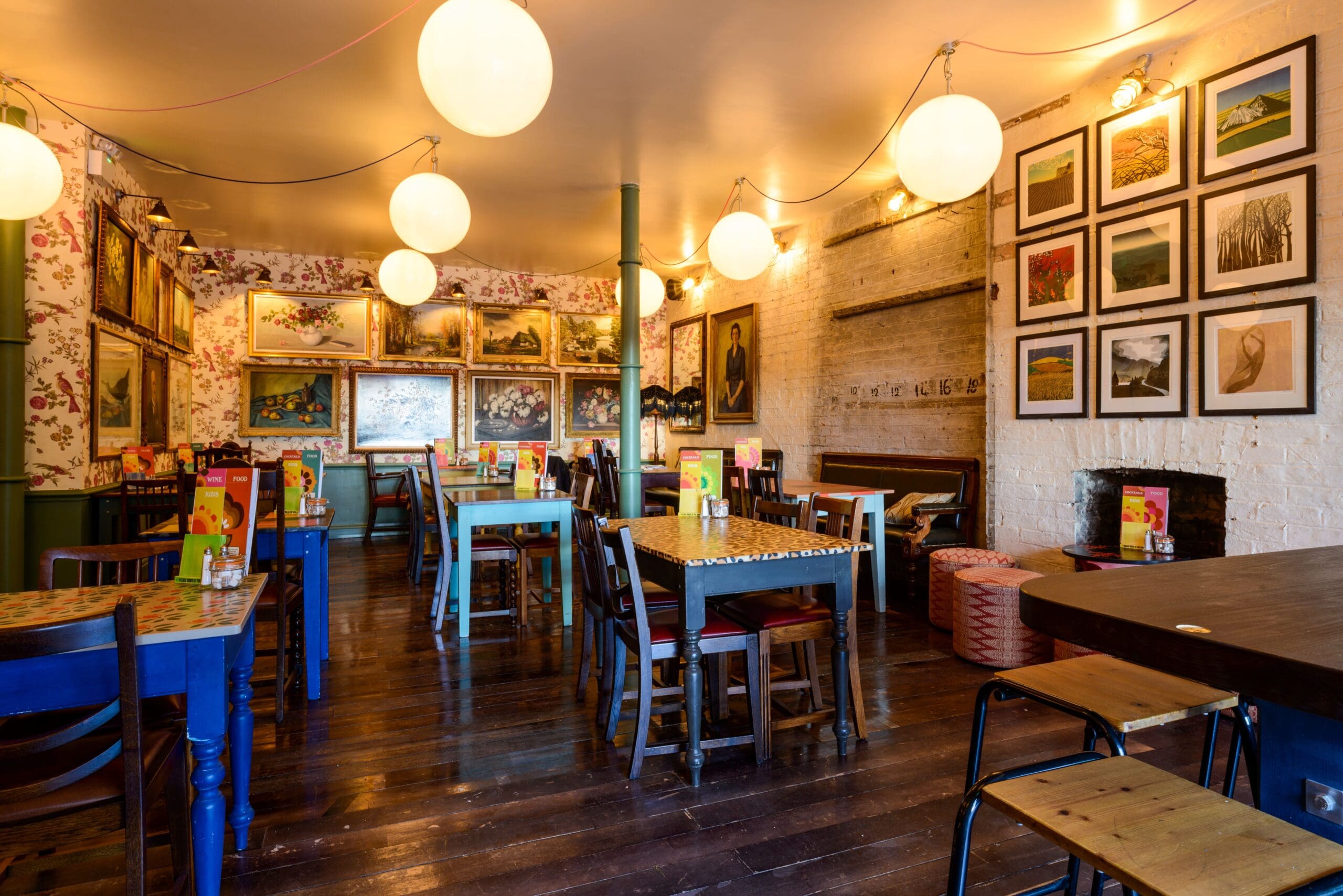 A cozy, brightly lit café with wooden floors, mismatched chairs, and colorful Montero tables. The walls are decorated with framed artwork and paintings. Round pendant lights hang from the ceiling, creating a warm atmosphere.