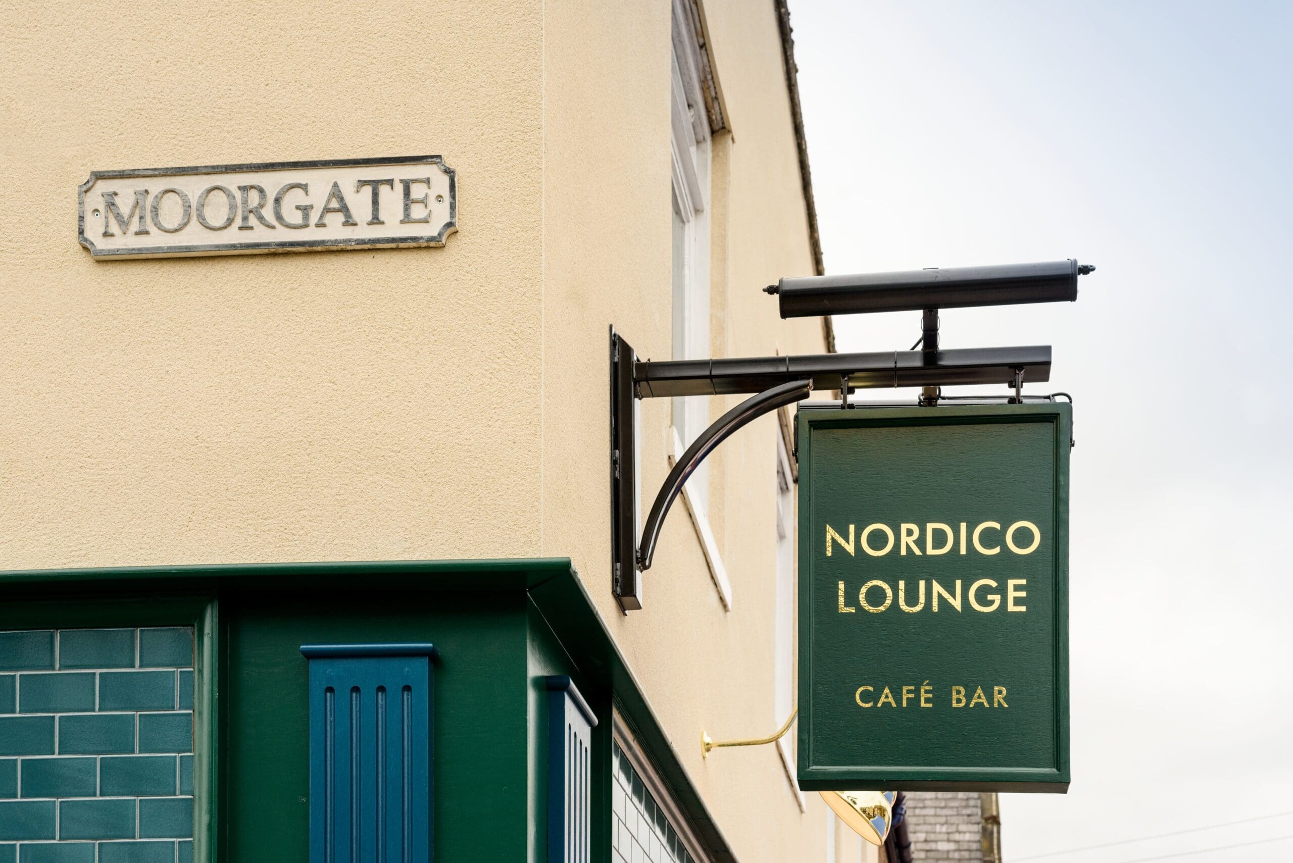 A green and gold Nordico Lounge Café Bar sign hangs on the side of a building with light-colored walls and green tile trim, just beneath a Moorgate street sign.