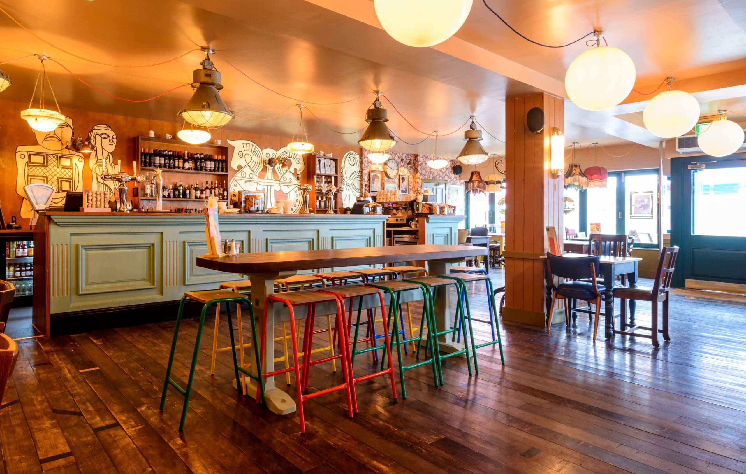 A cozy, warmly lit Nordico bar with colorful stools around a tall wooden table, wooden floors, and a decorated bar counter in the background. Hanging lights and quirky wall art create a welcoming, artistic atmosphere.