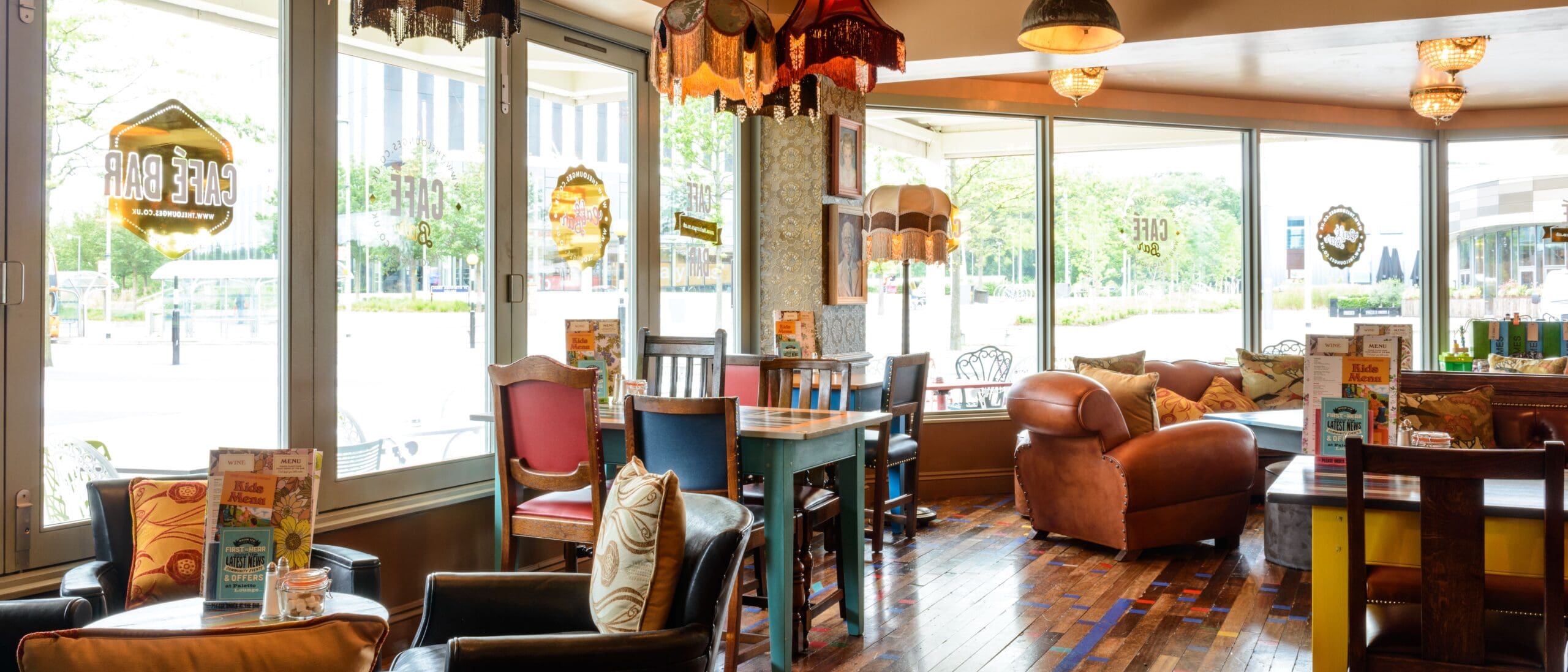 A cozy, sunlit café bar named Pablo with mismatched chairs, wooden tables, leather armchairs, and vintage lamps. Large windows fill Pablo’s space with natural light and offer charming views of trees and city buildings outside.