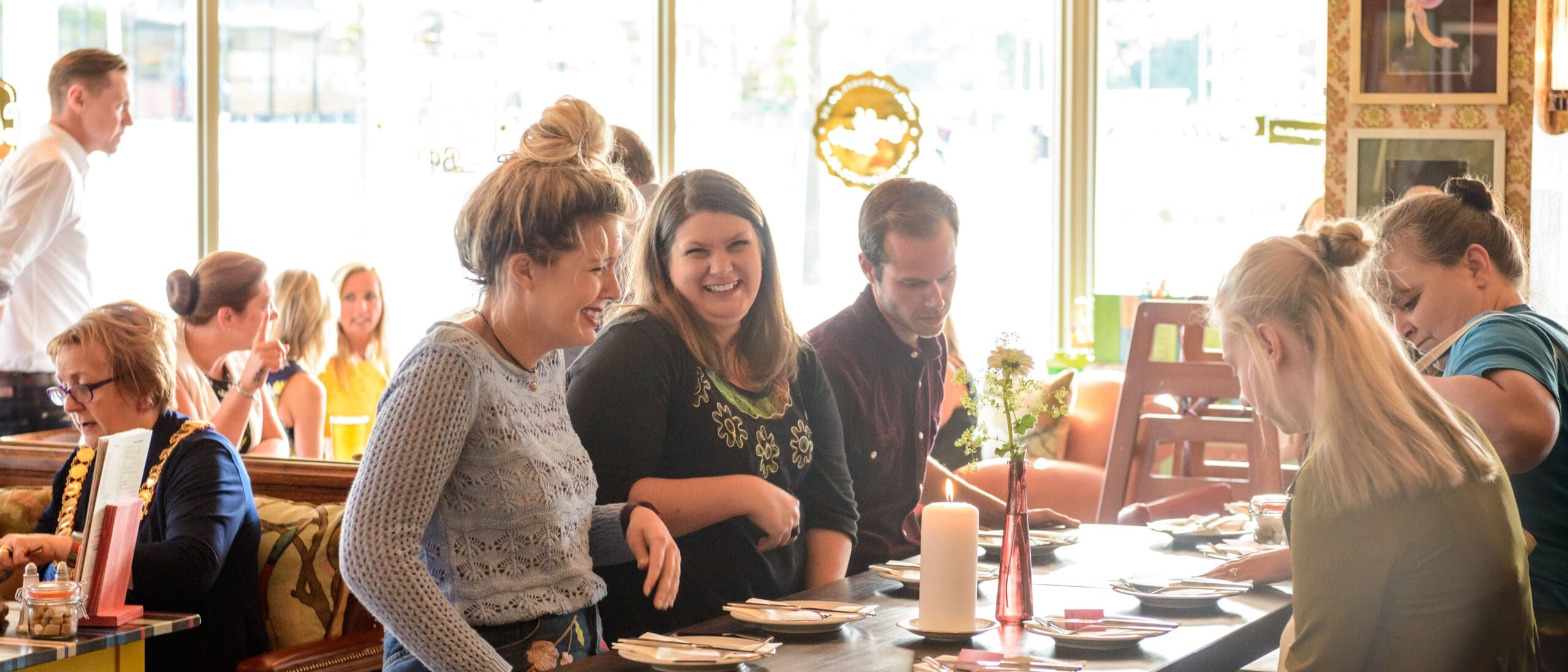 A group of people, including Pablo, sit and chat around a table at a lively, sunlit restaurant. Some are smiling and laughing, while others look at menus or arrange items on the table.