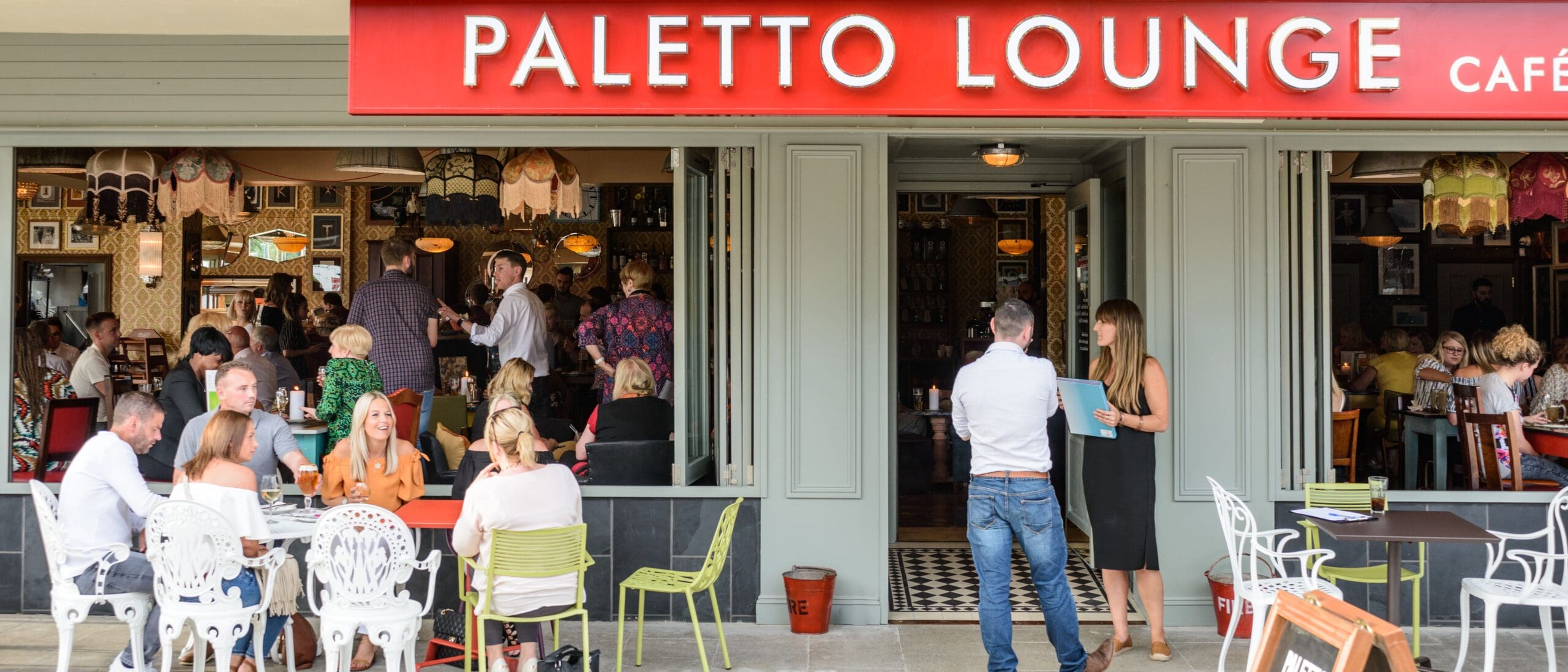 A busy café named Paletto Lounge with people sitting at outdoor tables, chatting and eating, while others are inside. At the entrance under the café's red sign, Pablo and a friend are deep in conversation.